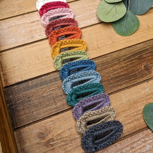 Puede incluir: Un conjunto de 14 pinzas para el pelo de crochet en varios colores, incluyendo blanco, rosa, rojo, naranja, amarillo, verde, azul, morado y gris. Las pinzas est&aacute;n dispuestas en una fila sobre una superficie de madera.