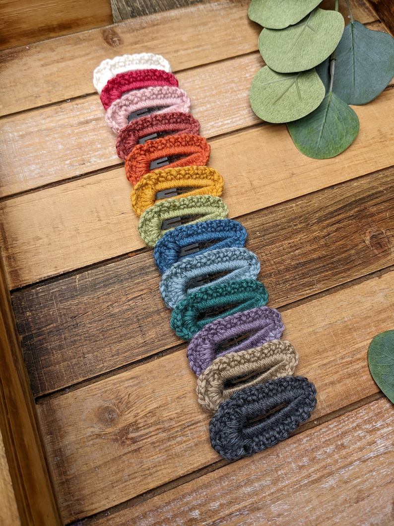 Puede incluir: Un conjunto de 14 pinzas para el pelo de crochet en varios colores, incluyendo blanco, rosa, rojo, naranja, amarillo, verde, azul, morado y gris. Las pinzas est&aacute;n dispuestas en una fila sobre una superficie de madera.