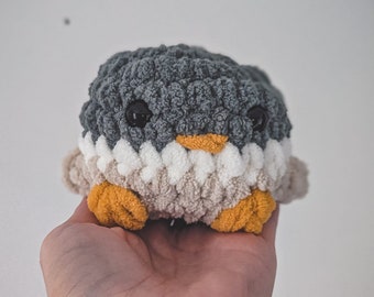 May include: A handmade, plush penguin toy. The penguin features a gray head, white and tan body, and orange feet. It has black button eyes and an orange beak. The toy is made of soft, textured yarn.