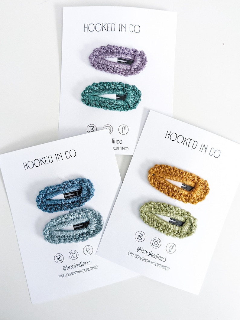 Puede incluir: Tres juegos de pinzas para el pelo de crochet en tarjetas blancas. Cada juego incluye dos pinzas en diferentes tonos de azul, verde y amarillo. Las tarjetas tienen el texto "HOOKED IN CO" y las cuentas de redes sociales.