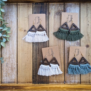 Puede incluir: Cuatro pares de pendientes en forma de triángulo con marcos de madera y borlas de flecos. Los pendientes se muestran sobre un fondo de madera. Las borlas son de color crema, verde y azul.