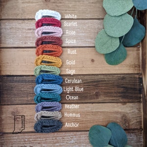 Puede incluir: Un conjunto de 13 pinzas para el pelo de crochet en varios colores, incluyendo blanco, escarlata, rosa, especias, &oacute;xido, oro, salvia, azul cer&uacute;leo, azul claro, oc&eacute;ano, brezo, hummus y ancla. Las pinzas est&aacute;n dispuestas verticalmente sobre una superficie de madera.
