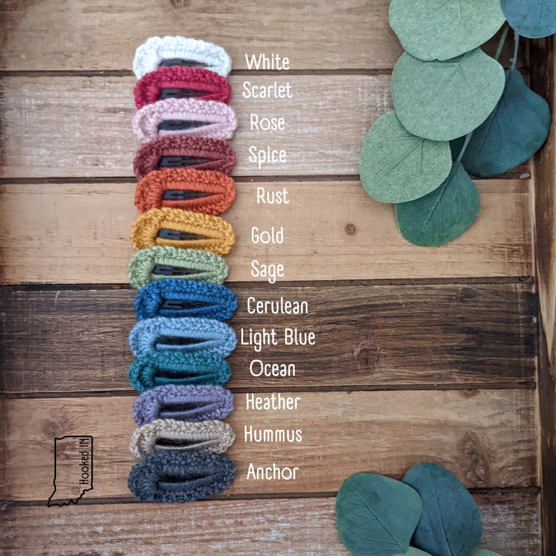Puede incluir: Un conjunto de 13 pinzas para el pelo de crochet en varios colores, incluyendo blanco, escarlata, rosa, especias, &oacute;xido, oro, salvia, azul cer&uacute;leo, azul claro, oc&eacute;ano, brezo, hummus y ancla. Las pinzas est&aacute;n dispuestas verticalmente sobre una superficie de madera.