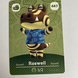 Peut inclure: Une carte Animal Crossing verte, marron et jaune représentant un personnage de crocodile nommé Roswell. Le numéro de la carte est 447 et le personnage porte une chemise bleue et blanche à motif ananas. La carte comprend également le texte "Roswell" et "5/2".