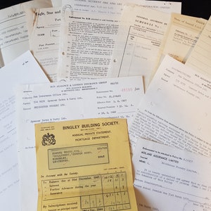 Op de afbeelding: Een verzameling vintage documenten, waaronder verzekeringspolissen en financi&euml;le overzichten, ligt verspreid. De papieren zijn verouderd met zichtbare kreukels en vervaagde inkt, wat duidt op historische waarde. De documenten bevatten tekst en diverse bedrijfslogo's.