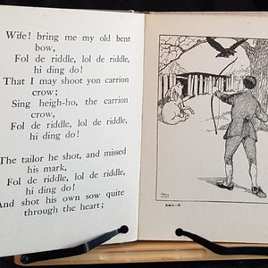 Op de afbeelding: Open boek met een zwart-wit illustratie van een man die met pijl en boog op een kraai schiet. De tekst op de linkerpagina begint met "Wife! bring me my old bent bow..." en gaat verder met een gedicht. Het boek wordt ondersteund door een zwarte standaard.