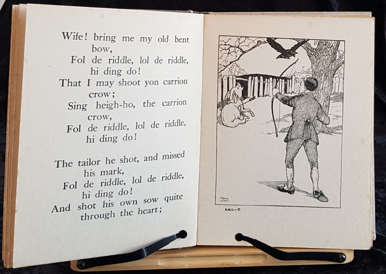 Op de afbeelding: Open boek met een zwart-wit illustratie van een man die met pijl en boog op een kraai schiet. De tekst op de linkerpagina begint met "Wife! bring me my old bent bow..." en gaat verder met een gedicht. Het boek wordt ondersteund door een zwarte standaard.