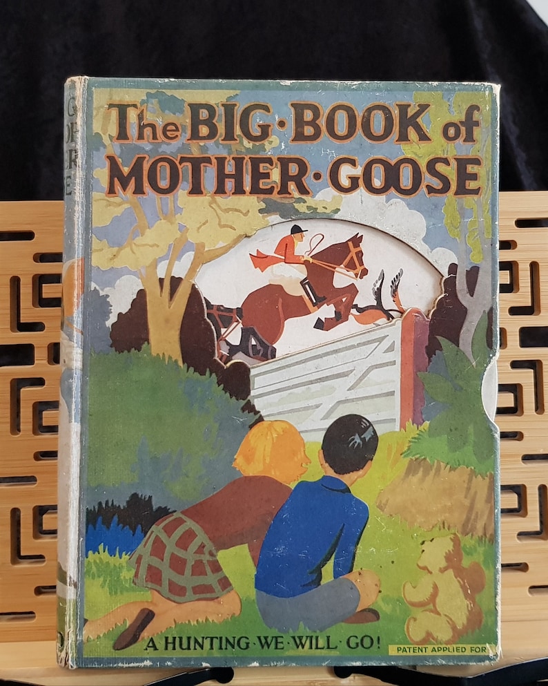 Op de afbeelding: Vintage kinderboek getiteld "The Big Book of Mother Goose". De omslag toont een kleurrijke illustratie van een paard dat over een hek springt, met twee kinderen die toekijken. De titel van het boek staat in grote, vette letters. De zin "A Hunting We Will Go!" staat onderaan.