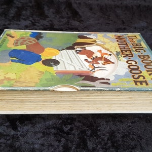 Op de afbeelding: Vintage kinderboek getiteld "The Big Book of Mother Goose". De omslag toont een kleurrijke illustratie van figuren en dieren in een landelijke sc&egrave;ne. De pagina's van het boek zijn verouderd en vertonen tekenen van slijtage.