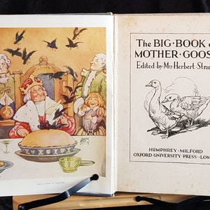 Op de afbeelding: Een open boek met kleurrijke illustraties en tekst. De linkerpagina toont een taart en figuren, terwijl de rechterpagina de titel "The Big Book of Mother Goose" en illustraties van ganzen weergeeft.