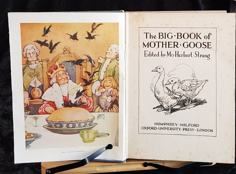 Op de afbeelding: Een open boek met kleurrijke illustraties en tekst. De linkerpagina toont een taart en figuren, terwijl de rechterpagina de titel "The Big Book of Mother Goose" en illustraties van ganzen weergeeft.