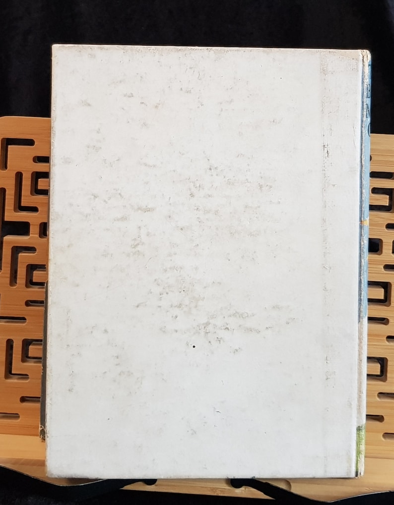 Op de afbeelding: Een vintage boek met een vervaagde witte kaft. De kaft vertoont tekenen van ouderdom met verkleuring en slijtage. De rug is zichtbaar met blauwe en groene accenten. Het boek ligt op een houten standaard met een geometrisch ontwerp.