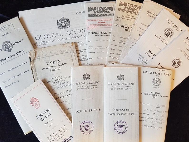 Op de afbeelding: Een verzameling vintage verzekeringsdocumenten, waaronder polissen en contracten, met diverse bedrijfslogo's en tekst. De documenten zijn in tinten wit en beige, met zwarte tekst en enkele rode accenten. Gerangschikt op een donkere ondergrond.
