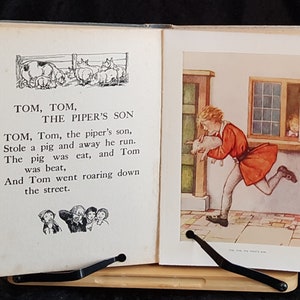 Op de afbeelding: Een opengevouwen vintage boek met illustraties en tekst. De linkerpagina toont een zwart-wit tekening van varkens en de tekst "Tom, Tom, The Piper's Son". De rechterpagina toont een jongen die met een varken rent.