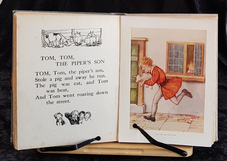 Op de afbeelding: Een opengevouwen vintage boek met illustraties en tekst. De linkerpagina toont een zwart-wit tekening van varkens en de tekst "Tom, Tom, The Piper's Son". De rechterpagina toont een jongen die met een varken rent.