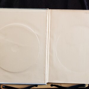 Op de afbeelding: Open boek met lichtgekleurde pagina's en cirkelvormige inkepingen. De tekst "1933 - 1967" is zichtbaar in de rechterbovenhoek. Het boek is open, met de binnenpagina's zichtbaar, en rust op een zwarte standaard.