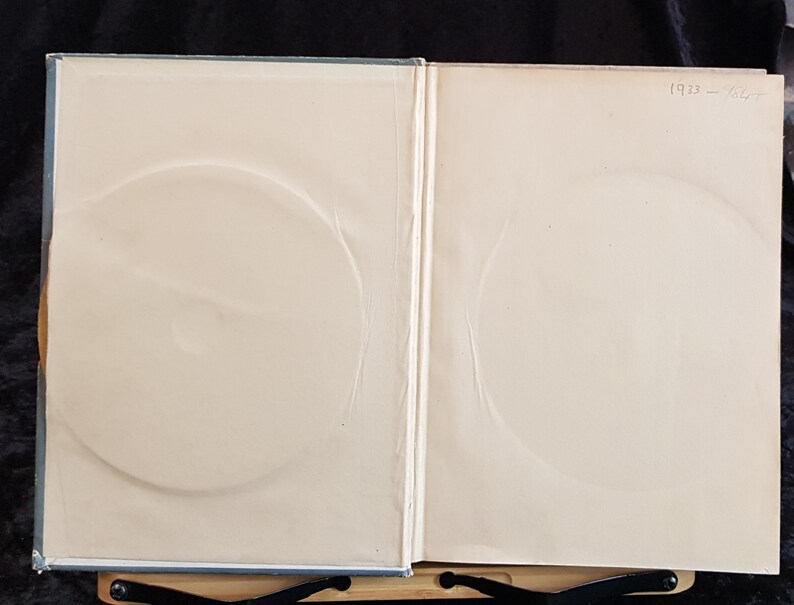 Op de afbeelding: Open boek met lichtgekleurde pagina's en cirkelvormige inkepingen. De tekst "1933 - 1967" is zichtbaar in de rechterbovenhoek. Het boek is open, met de binnenpagina's zichtbaar, en rust op een zwarte standaard.