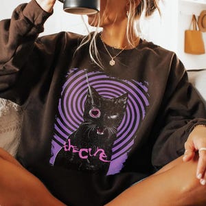 Puede incluir: Una sudadera marr&oacute;n oscuro con un gato negro, un ojo en forma de coraz&oacute;n y un dise&ntilde;o en espiral morado y blanco. El texto "the cure" est&aacute; impreso debajo del gato en cursiva.