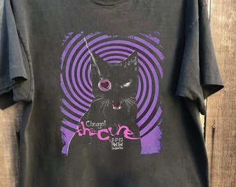 May include: Black t-shirt featuring a purple and black graphic of a cat with a heart-shaped eye in a spiral. The text "Chicago! The Cure 9.17-23 Red Star Douglas Park" is printed below the cat.