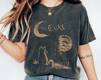 May include: A dark gray t-shirt with a vintage-style graphic of a crescent moon, a full moon, and a cat. The text "The Cure" is printed on the shirt.