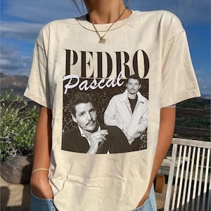 May include: Beige t-shirt featuring a black and white graphic of Pedro Pascal. The graphic includes the name "Pedro" in a bold font above the name "Pascal" in a script font, along with two images of the actor.
