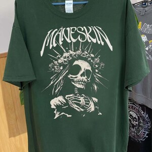 K&ouml;nnte beinhalten: Dunkelgr&uuml;nes T-Shirt mit dem Bandnamen "MANESKIN" in Wei&szlig;, &uuml;ber einer wei&szlig;en Grafik eines Sch&auml;dels mit einem Totenkronen-Kopfschmuck und einem Skelettorso. Das T-Shirt h&auml;ngt an einem Kleiderb&uuml;gel.