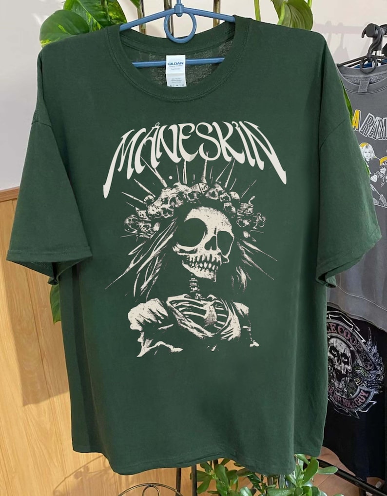 K&ouml;nnte beinhalten: Dunkelgr&uuml;nes T-Shirt mit dem Bandnamen "MANESKIN" in Wei&szlig;, &uuml;ber einer wei&szlig;en Grafik eines Sch&auml;dels mit einem Totenkronen-Kopfschmuck und einem Skelettorso. Das T-Shirt h&auml;ngt an einem Kleiderb&uuml;gel.