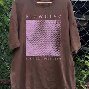 May include: A brown t-shirt with a pink and white graphic print of a face and the text "slowdive souvlaki tour 1994".