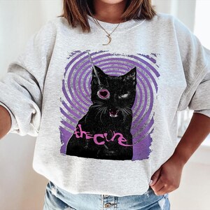 Puede incluir: Sudadera gris claro con un gr&aacute;fico de gato negro, un ojo de coraz&oacute;n rosa y el texto "the cure" en rosa. El gato est&aacute; sobre un fondo de espiral p&uacute;rpura.