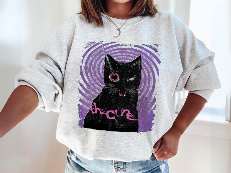 Puede incluir: Sudadera gris claro con un gr&aacute;fico de gato negro, un ojo de coraz&oacute;n rosa y el texto "the cure" en rosa. El gato est&aacute; sobre un fondo de espiral p&uacute;rpura.