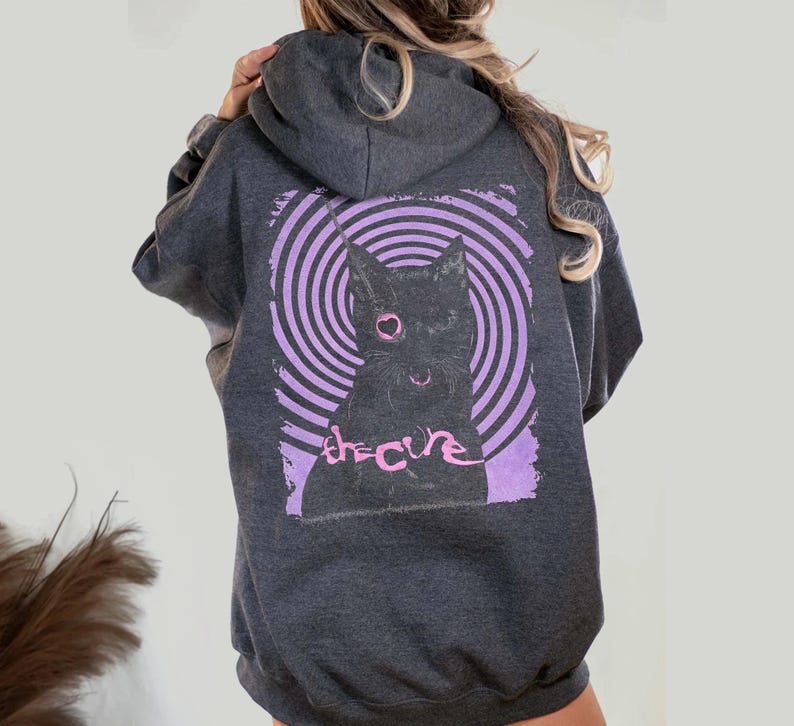 Puede incluir: Sudadera con capucha gris oscuro con una ilustraci&oacute;n de un gato negro con un ojo y una nariz en forma de coraz&oacute;n rosa, sobre un fondo de espiral p&uacute;rpura. El texto "the cure" est&aacute; impreso en rosa. La sudadera tiene una capucha.