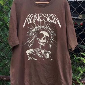 K&ouml;nnte beinhalten: Braunes T-Shirt mit dem Bandnamen "Maneskin" in Wei&szlig;, &uuml;ber einer detaillierten Skelettgrafik. Das Skelett tr&auml;gt eine Krone und florale Elemente. Das T-Shirt h&auml;ngt an einem Holzb&uuml;gel.