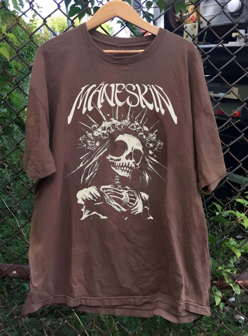 K&ouml;nnte beinhalten: Braunes T-Shirt mit dem Bandnamen "Maneskin" in Wei&szlig;, &uuml;ber einer detaillierten Skelettgrafik. Das Skelett tr&auml;gt eine Krone und florale Elemente. Das T-Shirt h&auml;ngt an einem Holzb&uuml;gel.