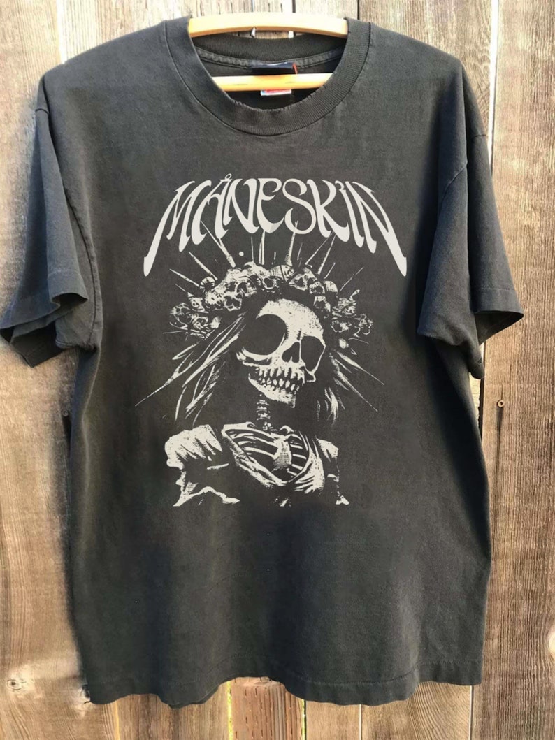 K&ouml;nnte beinhalten: Dunkelgraues T-Shirt mit dem Bandnamen "MANESKIN" in Wei&szlig; &uuml;ber einer wei&szlig;en Grafik eines Sch&auml;dels, der eine Krone aus Totenk&ouml;pfen tr&auml;gt. Das Design umfasst einen Skelettorso und strahlenf&ouml;rmige Linien.