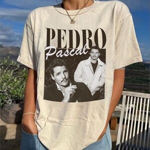 May include: Beige t-shirt featuring a black and white image of Pedro Pascal with the text "PEDRO Pascal" in a vintage style. The graphic includes two images of the actor. The shirt is a neutral color and has short sleeves.