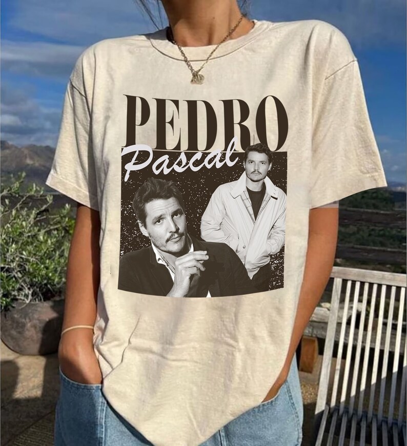 May include: Beige t-shirt featuring a black and white image of Pedro Pascal with the text "PEDRO Pascal" in a vintage style. The graphic includes two images of the actor. The shirt is a neutral color and has short sleeves.