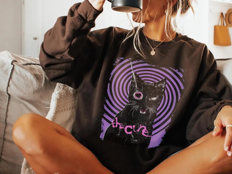 Puede incluir: Una sudadera marr&oacute;n oscuro con un gato negro, un ojo en forma de coraz&oacute;n y un dise&ntilde;o en espiral morado y blanco. El texto "the cure" est&aacute; impreso debajo del gato en cursiva.