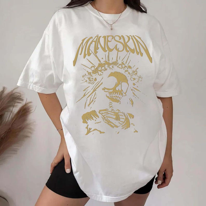 K&ouml;nnte beinhalten: Wei&szlig;es T-Shirt mit dem Bandnamen "Maneskin" in goldener Schrift &uuml;ber einer goldenen Skelettgrafik. Das kurz&auml;rmelige T-Shirt hat einen Rundhalsausschnitt. Das Design ist mittig auf der Vorderseite platziert.