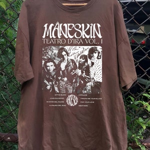 May include: Brown t-shirt with a white print of the band M&aring;neskin. The print includes the band's name, the album title "Teatro d'Ira Vol. 1", and a photo of the band members. The album title is written in a cursive font. The band members are pictured in black and white. The t-shirt also includes a list of the songs on the album.