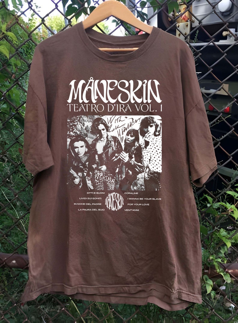 May include: Brown t-shirt with a white print of the band M&aring;neskin. The print includes the band's name, the album title "Teatro d'Ira Vol. 1", and a photo of the band members. The album title is written in a cursive font. The band members are pictured in black and white. The t-shirt also includes a list of the songs on the album.