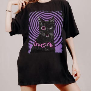 Puede incluir: Camiseta negra con un gr&aacute;fico de un gato negro con un ojo en forma de coraz&oacute;n y el texto "the cure" en rosa. El gato est&aacute; sobre un fondo de espiral morado y negro. La camiseta tiene mangas cortas y cuello redondo.