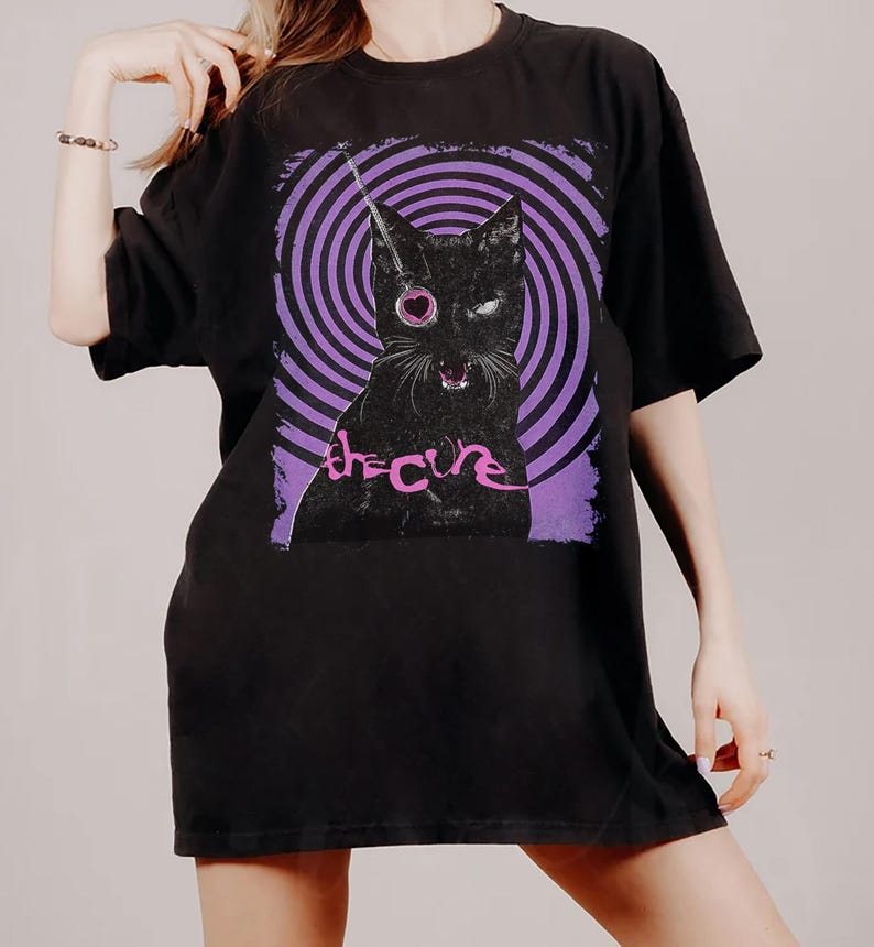 Puede incluir: Camiseta negra con un gr&aacute;fico de un gato negro con un ojo en forma de coraz&oacute;n y el texto "the cure" en rosa. El gato est&aacute; sobre un fondo de espiral morado y negro. La camiseta tiene mangas cortas y cuello redondo.