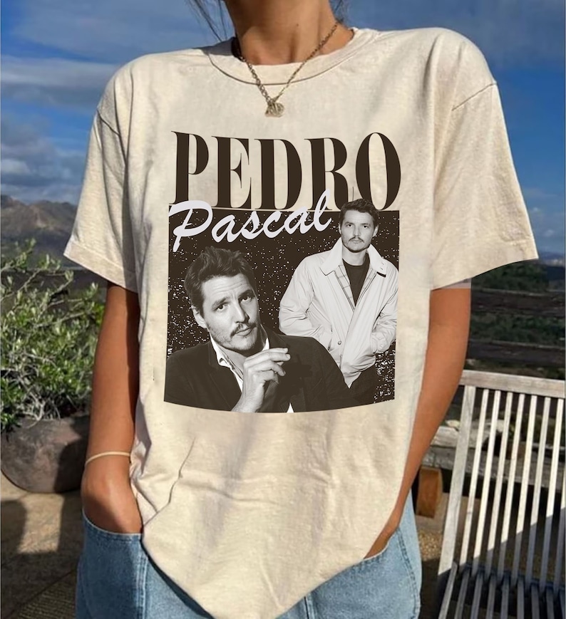 May include: Beige t-shirt featuring a black and white graphic of Pedro Pascal. The graphic includes the name "Pedro" in a bold font above the name "Pascal" in a script font, along with two images of the actor.