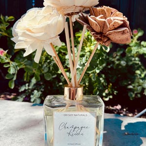 May include: A clear glass oil reed diffuser with a gold-colored collar, containing a light-colored liquid. The label reads "Champagne Kisses" and "Oh Soy Naturals." The diffuser is topped with decorative wooden flowers and reeds.