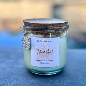 May include: A clear glass candle jar with a cork lid, tied with twine and a small silver charm. The white label features a pink starfish graphic, the text Oh Soy Naturals, Island Girl, Hand Poured in Galt, CA, and the weight 10 Oz (284 g).