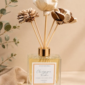 May include: A square glass reed diffuser bottle filled with light yellow liquid, topped with a gold collar and three wooden sticks featuring decorative sola wood flowers. The white label reads OH SOY NATURALS, Champagne Kisses, champagne, citrus, plumeria, jasmine, Oil Reed Diffuser, 5 oz (141g).