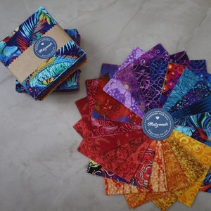 May include: A selection of patterned fabric squares, some neatly stacked and bound, others fanned out to display their designs. The fabrics showcase vivid patterns in blue, red, orange, and purple. A blue label with the text "Matzmade" is visible.