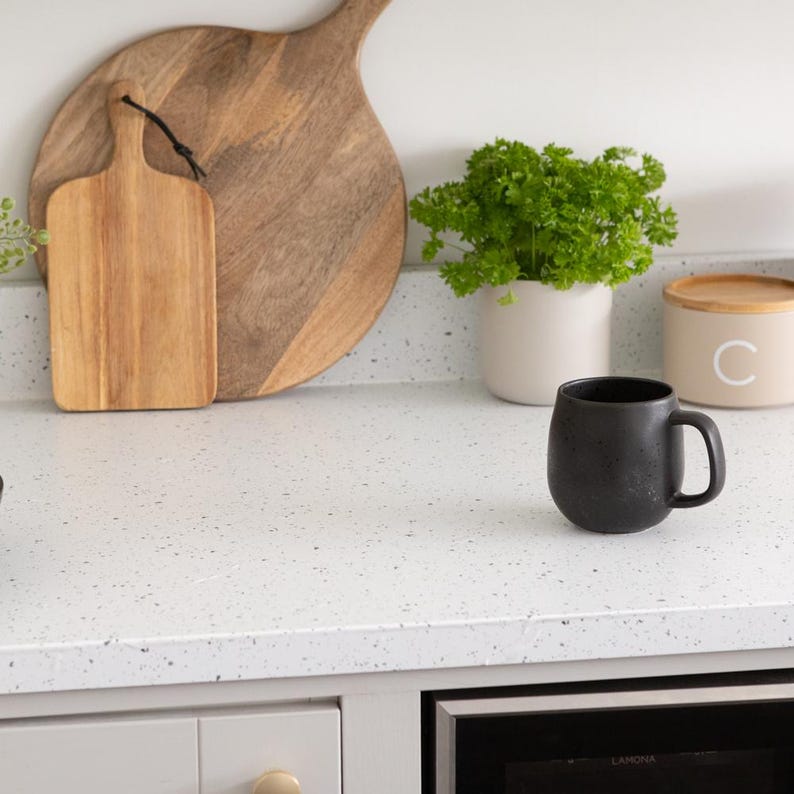 May include: A kitchen countertop scene featuring a black mug, wooden cutting boards, a small potted plant, and a canister with a letter "C". The countertop is white with speckled details, and the overall aesthetic is modern and minimalist.