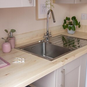May include: A stainless steel kitchen sink with a chrome faucet is set into a light wood countertop. A pink soap dispenser and vase are on the counter, along with a small plant in a white pot. The cabinets are light gray.
