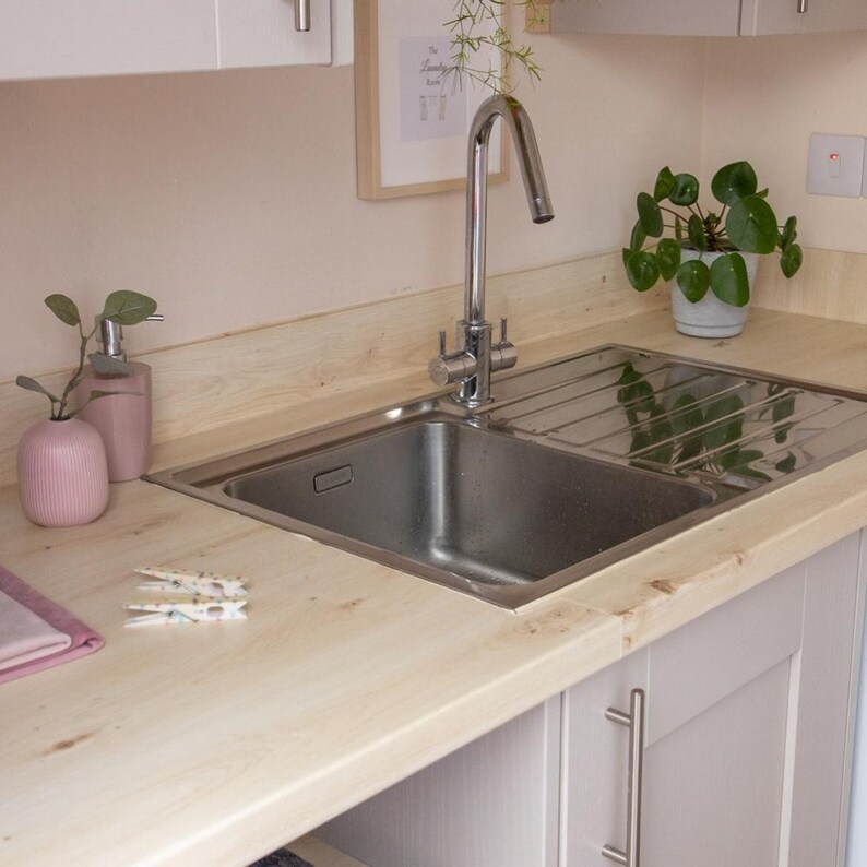 May include: A stainless steel kitchen sink with a chrome faucet is set into a light wood countertop. A pink soap dispenser and vase are on the counter, along with a small plant in a white pot. The cabinets are light gray.